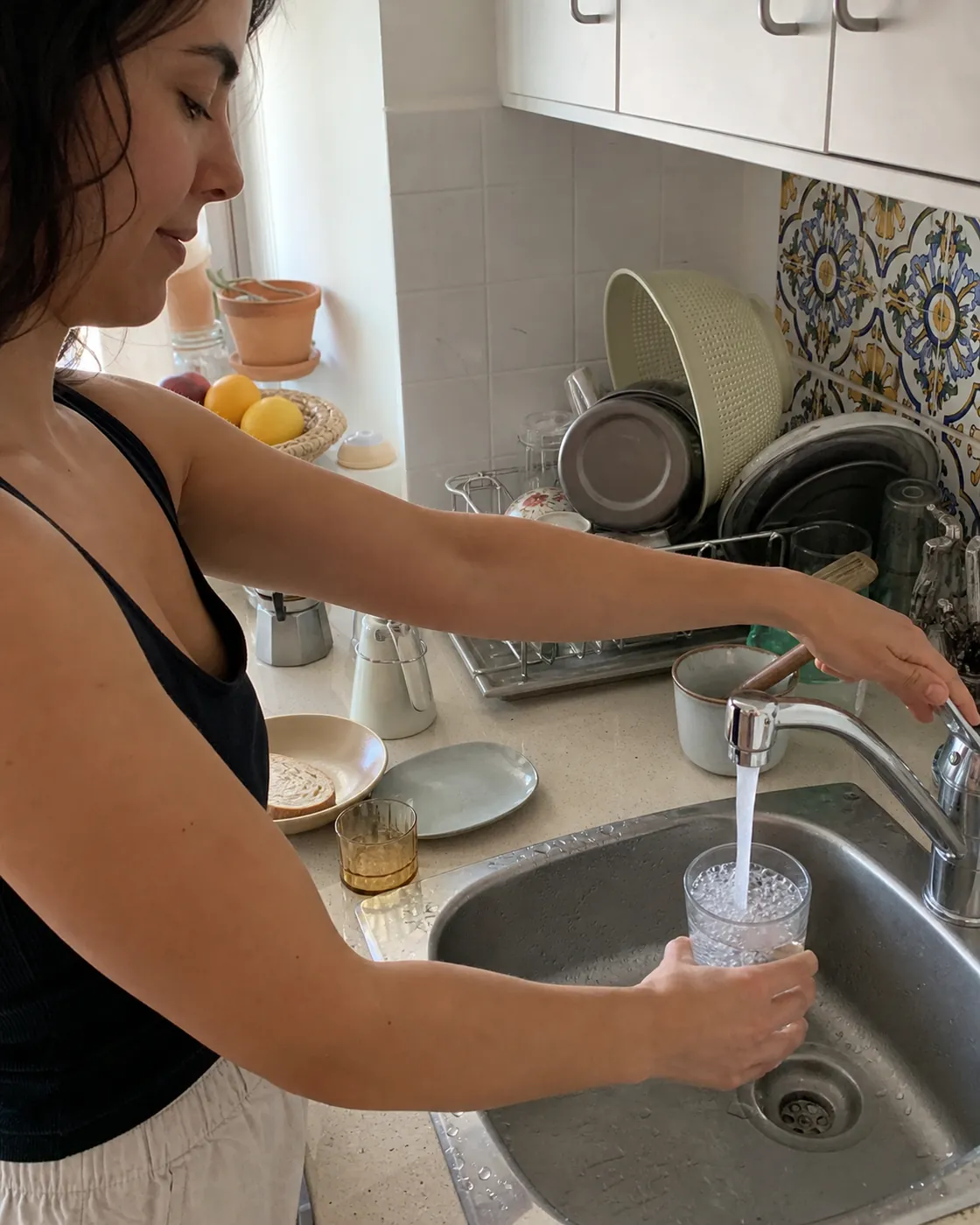 A woman filling a glass of tap water at the sink in a tiled Valencian kitchen